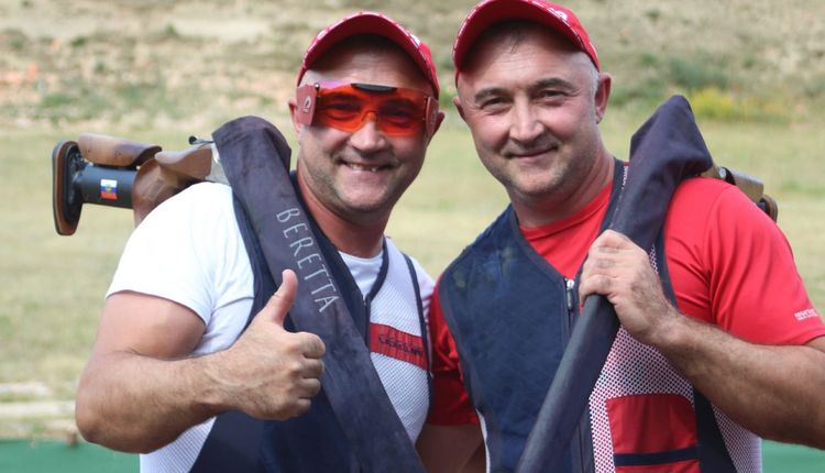 Zwei Flinten-Schützen blicken in die Kamera und machen die "Daumen hoch" Geste. Ihre Sportgeräte haben sie über die Schultern gelegt.