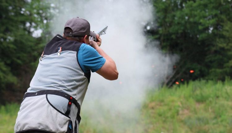 Ein Flintenschütze im Wettkampf. Pulverdampf, im Hintergrund eine zerspringende Wurfscheibe.